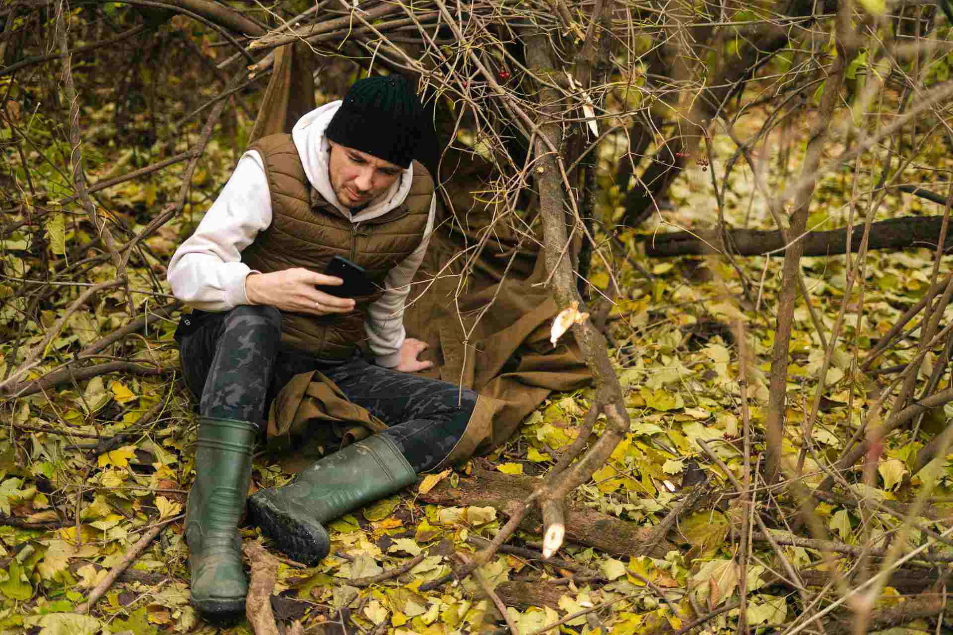 Man in a forest shelter using a smartphone, dressed in outdoor gear and rubber boots.