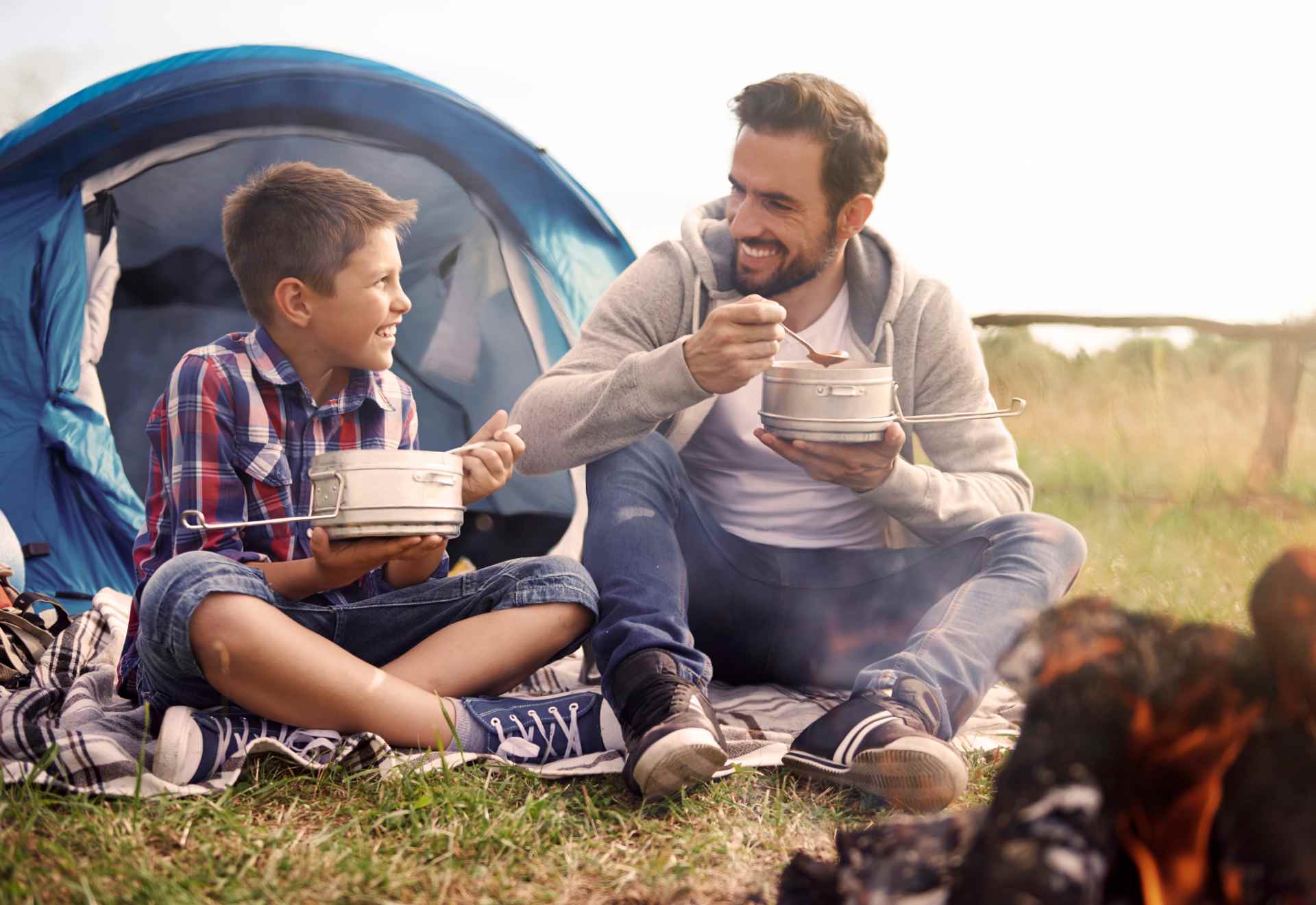 A father and son sitting outdoors near a blue camping tent, smiling and enjoying a meal from camping pots. A campfire is visible in the foreground.