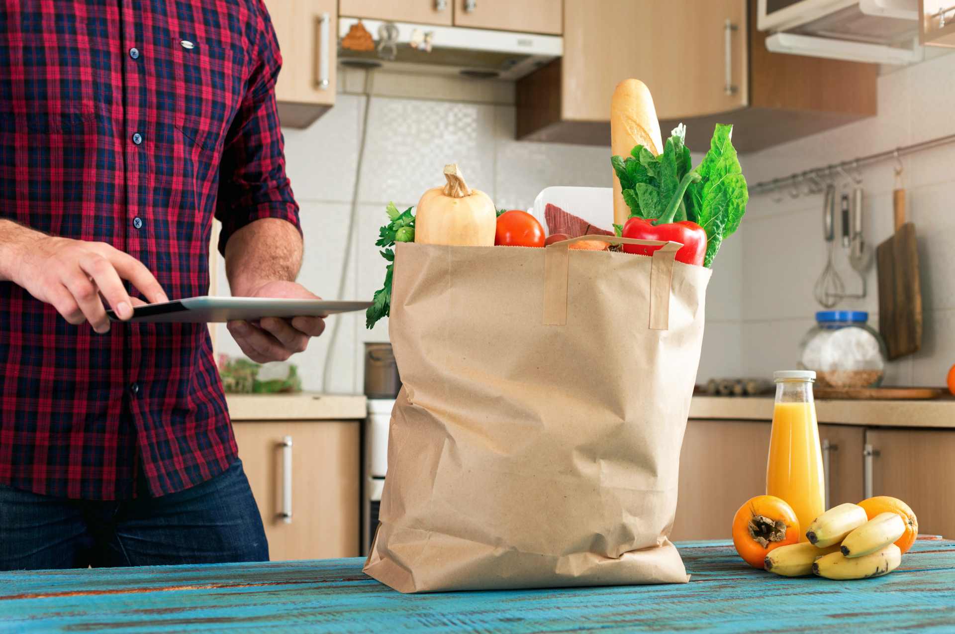 A person using a tablet near a grocery bag with fresh produce on a kitchen counter.
