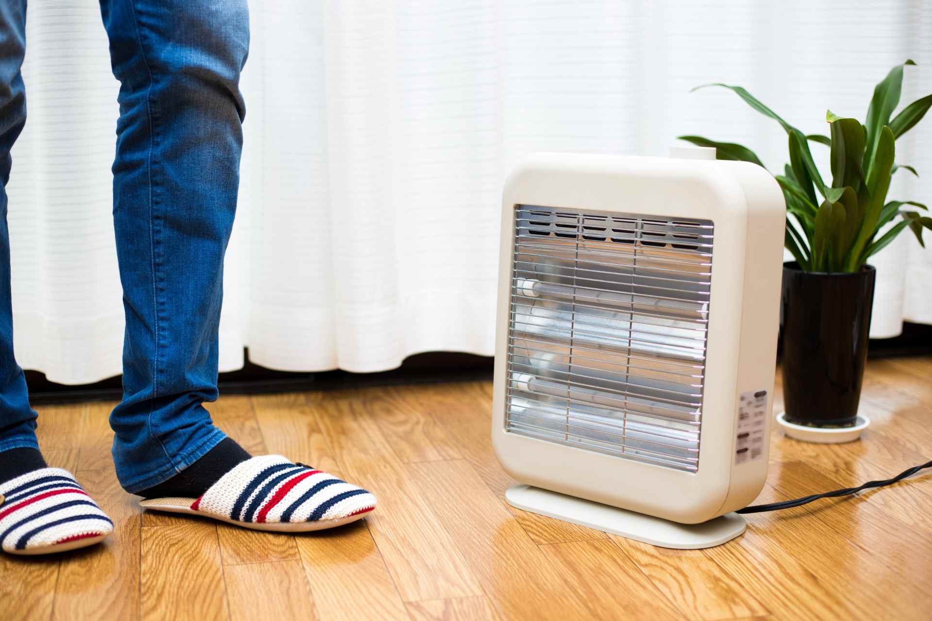 A small white electric heater operating on a wooden floor with striped slippers and a plant in the background, showing the contrast between different types of heaters.
