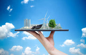 A hand holding a miniature 3D model of a sustainable city with wind turbines, solar panels, and trees against a blue sky backdrop, symbolizing the concept of green energy and self-sufficient, eco-friendly living.
