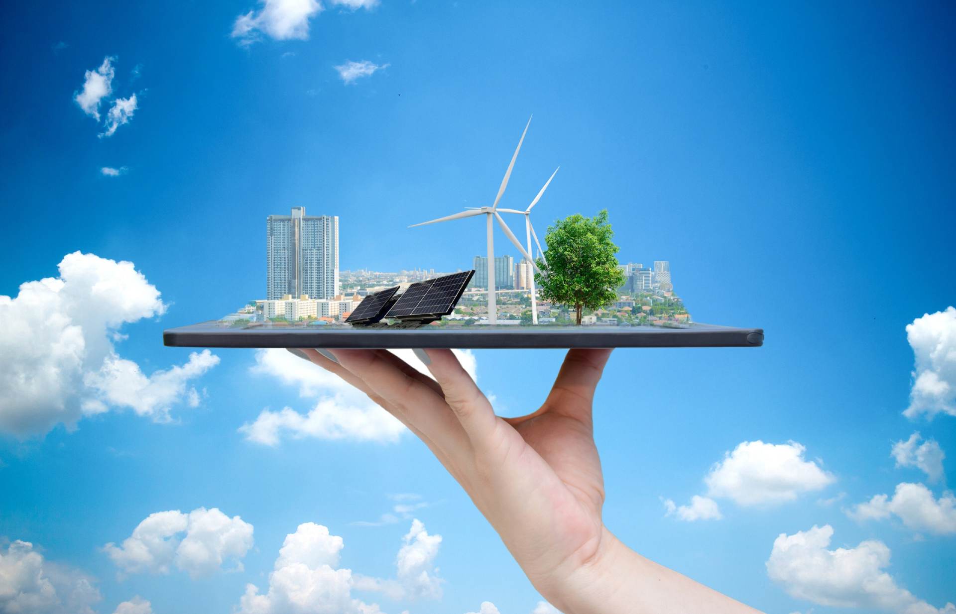 A hand holding a miniature 3D model of a sustainable city with wind turbines, solar panels, and trees against a blue sky backdrop, symbolizing the concept of green energy and self-sufficient, eco-friendly living.