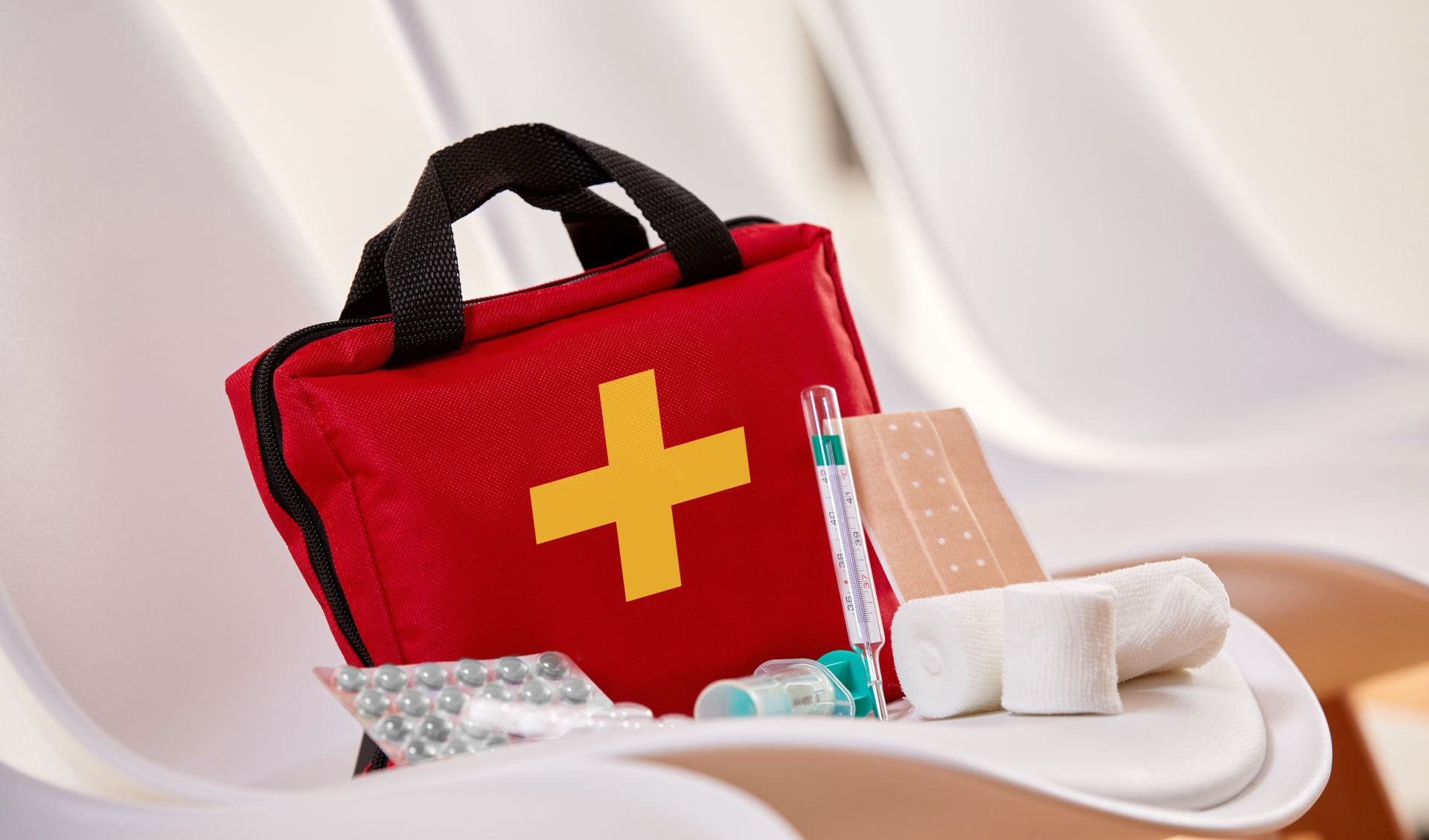 A red first aid kit with a bright yellow cross on the front, placed on a white surface, accompanied by various medical supplies, including gauze, bandages, pills, and a thermometer.