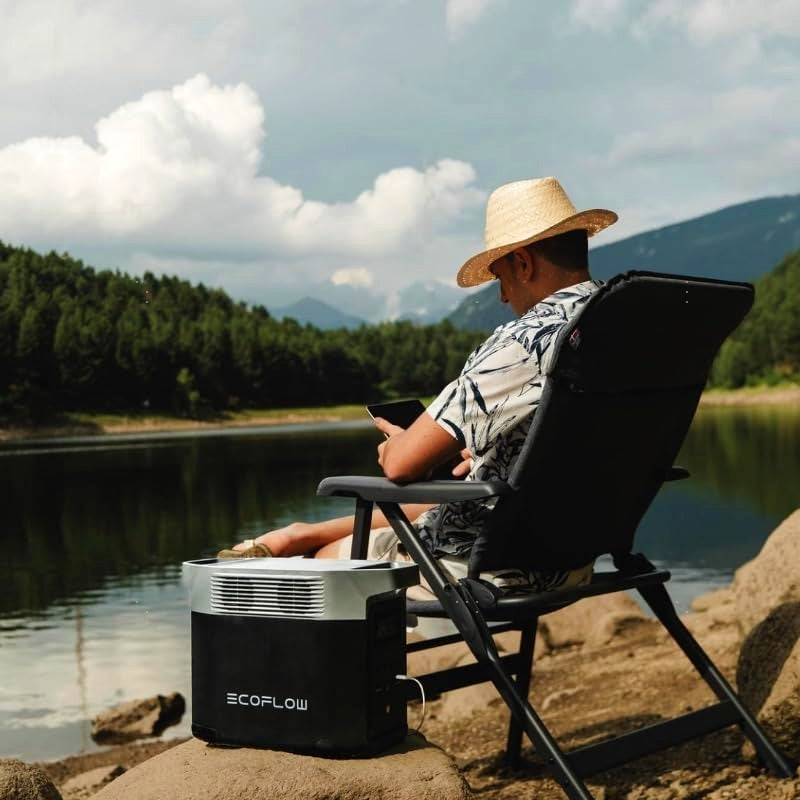 A man relaxing in a chair near a serene lake, using his phone while an EcoFlow portable power station sits beside him, highlighting its usability in outdoor adventures.