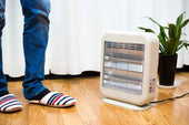 A small white electric heater operating on a wooden floor with striped slippers and a plant in the background, showing the contrast between different types of heaters.