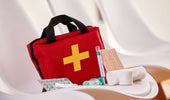  A red first aid kit with a bright yellow cross on the front, placed on a white surface, accompanied by various medical supplies, including gauze, bandages, pills, and a thermometer.
